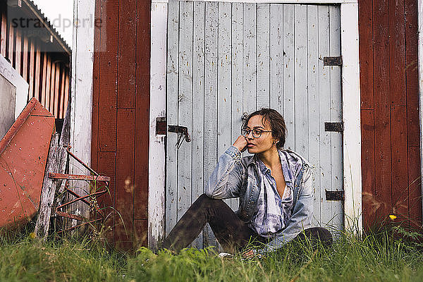 Porträt einer jungen Frau  die im Gras vor einer Holzhütte sitzt