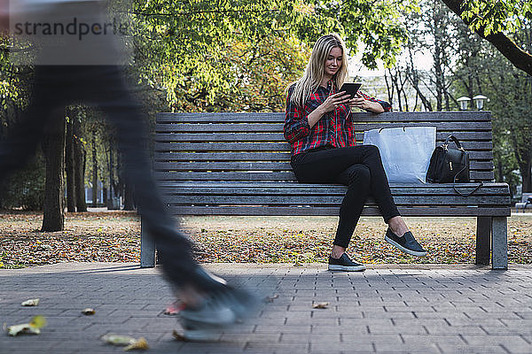 Junge Frau mit Einkaufstasche sitzt im Herbst mit Minitablette auf Bank