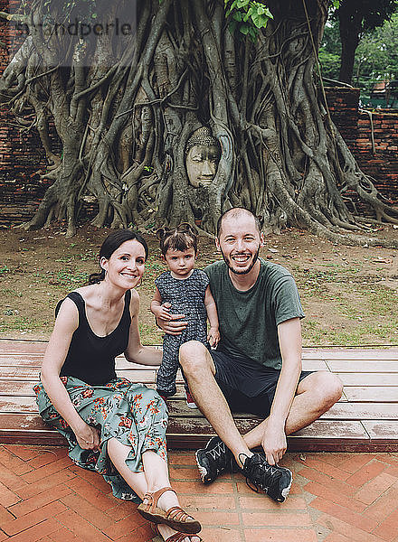 Thailand  Ayutthaya  Porträt einer lächelnden Familie  die den Buddhakopf zwischen Baumwurzeln im Wat Mahathat besucht