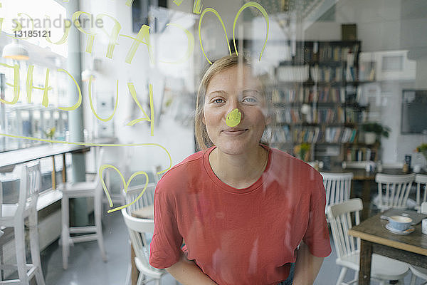 Porträt einer lächelnden jungen Frau  die hinter einer Fensterscheibe in einem Cafe posiert