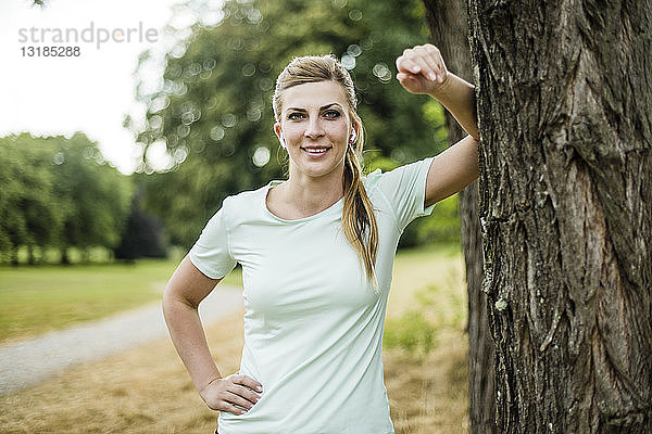 Lächelnde  sportliche junge Frau  die sich an einen Baum in einem Park lehnt