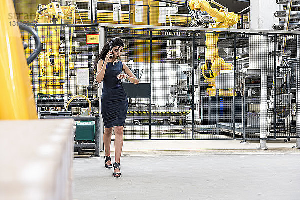 Frau am Telefon geht in Fabrikhalle mit Industrieroboter  der die Zeit überprüft