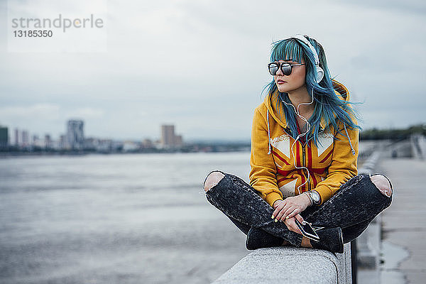 Junge Frau mit blau gefärbten Haaren sitzt an einer Wand und hört mit Kopfhörern und Smartphone Musik