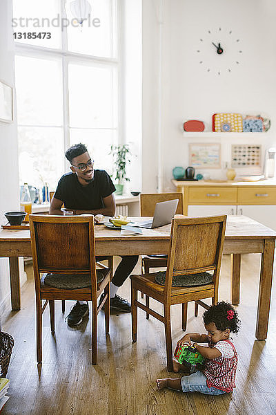 Kleines Mädchen spielt mit Spielzeugauto  während der Vater im Haus am Esstisch sitzt