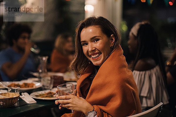 Porträt einer lächelnden jungen Frau  die während einer Dinnerparty im Hinterhof ein Getränk in der Hand hält  während sie am Tisch sitzt