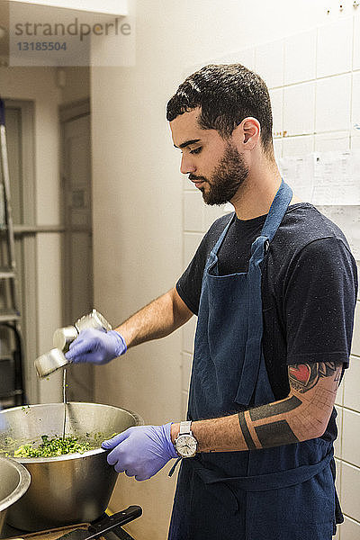 Selbstbewusster junger männlicher Koch  der in der Großküche eines Restaurants Speisen in einem Behälter zubereitet