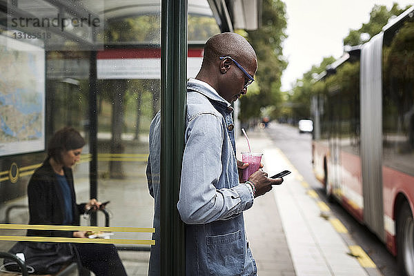 Geschäftsmann benutzt Smartphone  während er ein Getränk an einer Bushaltestelle in der Stadt hält