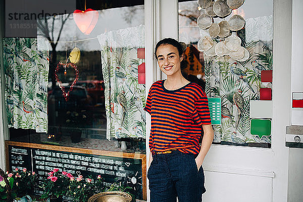 Porträt einer lächelnden jungen Frau  die mit der Hand in der Tasche vor der Restauranttür steht