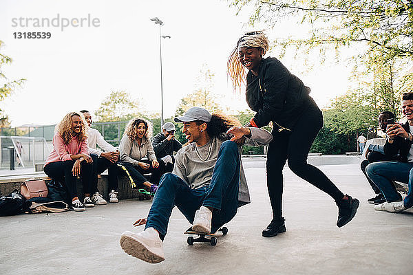 Teenager drängt jungen Mann auf Skateboard  während Freunde im Park lachen