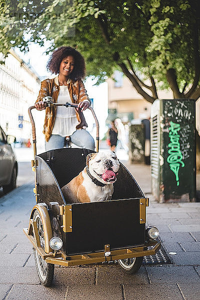 Lächelnde Frau fährt Fahrradwagen mit Bulldogge auf gepflasterter Straße