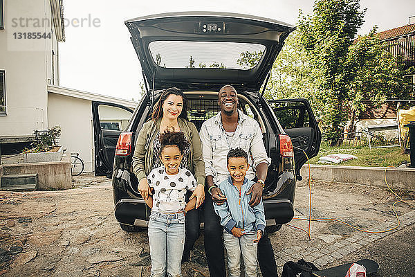 Porträt einer lächelnden multiethnischen Familie  die sich auf den Kofferraum eines Autos im Vorgarten lehnt