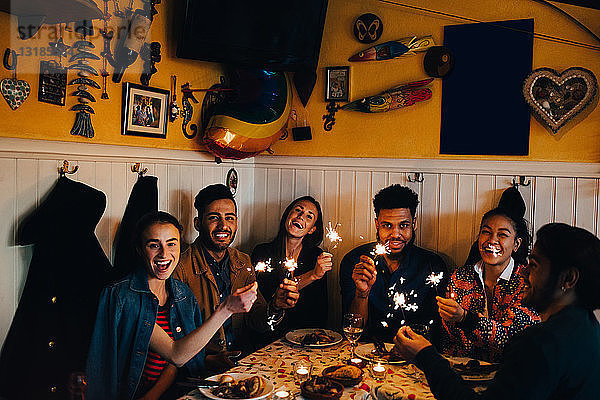 Porträt fröhlicher junger multi-ethnischer Freunde  die mit brennenden Wunderkerzen am Tisch im Restaurant sitzen