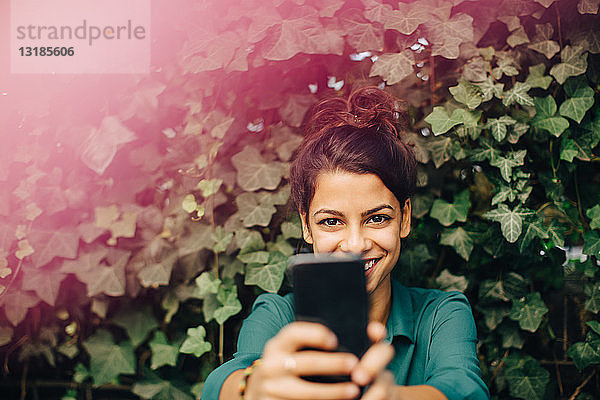Porträt einer lächelnden jungen Frau beim Fotografieren durch ein Mobiltelefon im Hinterhof
