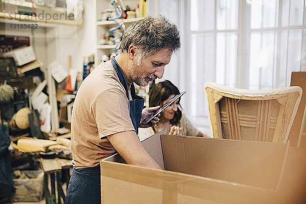 Männlicher Handwerker hält digitales Tablett in der Hand  während er in der Werkstatt in einen Karton schaut