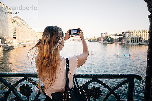 Rückansicht einer jungen Frau  die mit einem Mobiltelefon in der Stadt einen Fluss fotografiert