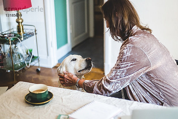 Frau streichelt Hund  während sie zu Hause Kaffee auf dem Esstisch trinkt