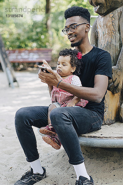 Kleinkind benutzt Mobiltelefon  während es mit seinem Vater auf einem Spielgerät im Freien auf dem Spielplatz sitzt