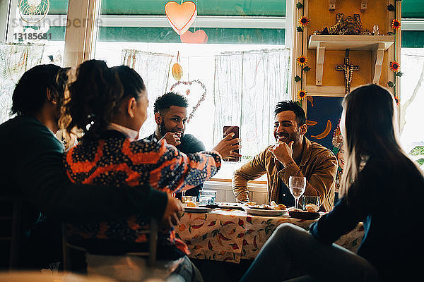 Junge Frau zeigt lächelndem Mann  der während der Brunch-Party im Restaurant sitzt  ihr Smartphone