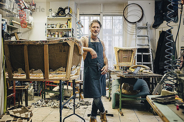 Porträt eines reifen männlichen Handwerkers neben einem unvollständigen Sofa in einer Polsterwerkstatt