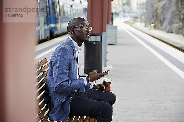 Nachdenklicher Geschäftsmann sitzt mit Smartphone und Kaffee am Bahnhof
