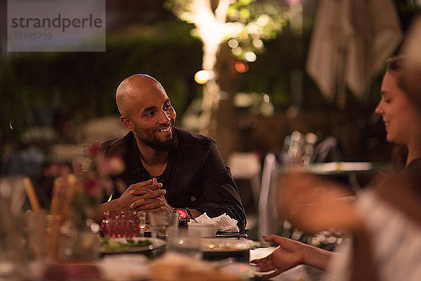 Lächelnder junger Mann mit rasiertem Kopf schaut eine Freundin an  während er während der Dinnerparty am Tisch sitzt