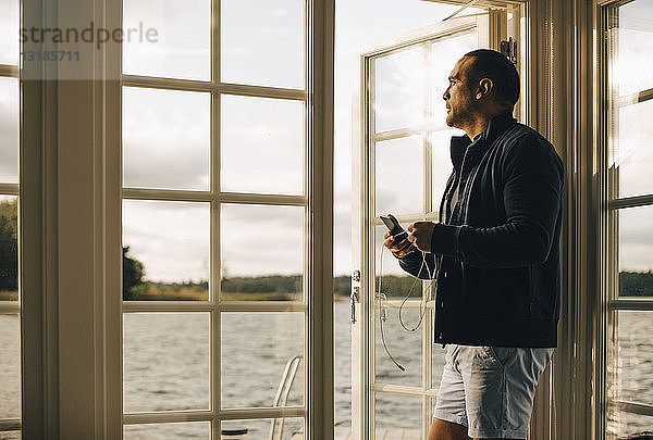 Nachdenklicher  reifer Mann benutzt Mobiltelefon  während er an der Türöffnung einer Ferienvilla steht