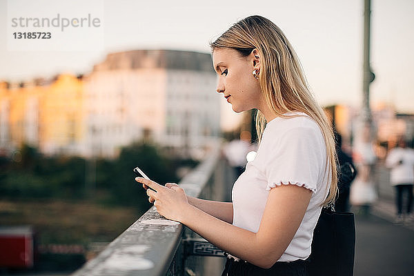 Seitenansicht einer jungen Frau  die ein Smartphone benutzt  während sie auf einer Brücke in der Stadt steht
