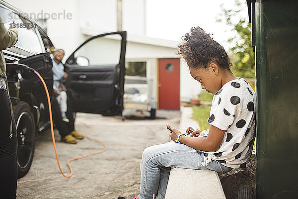 Seitenansicht eines Mädchens  das ein Smartphone benutzt  während es im Vorgarten sitzt