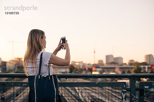 Junge Frau fotografiert durch ein Mobiltelefon  während sie bei Sonnenuntergang auf einer Brücke vor klarem Himmel steht