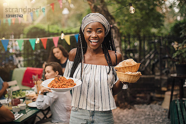 Porträt einer lächelnden jungen Frau  die Essen trägt  während sie während einer Gartenparty im Hinterhof steht