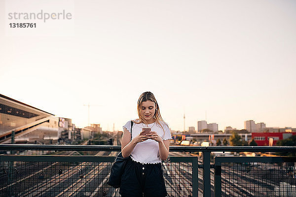 Junge Frau benutzt Mobiltelefon  während sie auf der Brücke gegen den klaren Himmel in der Stadt steht