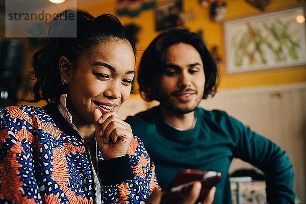 Lächelnder junger Mann und lächelnde Frau teilen sich ein Smartphone  während sie während des Brunch im Restaurant sitzen