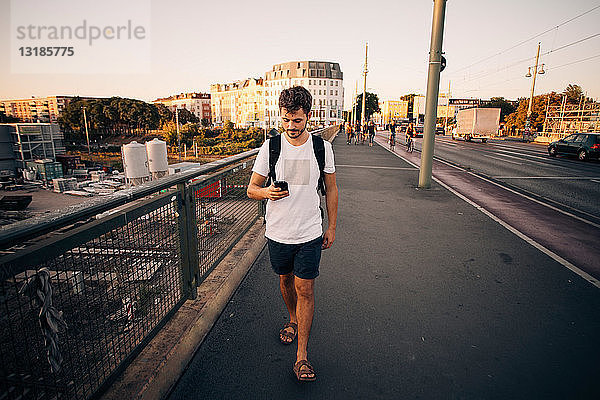 Ein junger Mann in voller Länge benutzt ein Mobiltelefon  während er auf einem Fußweg an einer Brücke in der Stadt läuft