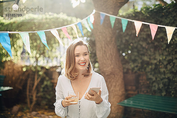Lächelnde junge Frau hält Getränk und Mobiltelefon in der Hand  während sie im Hinterhof steht