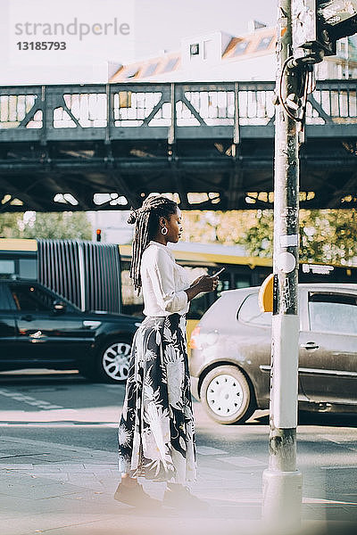 Seitenansicht einer jungen Frau  die ein Mobiltelefon benutzt  während sie in der Stadt auf dem Bürgersteig steht