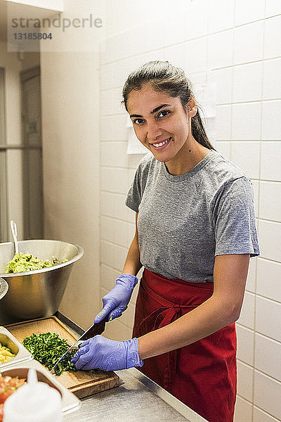 Lächelnder junger Chefkoch beim Zerkleinern von Gemüse auf dem Schneidebrett in der Großküche eines Restaurants