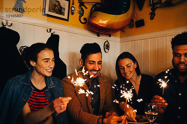 Glückliche junge multiethnische Freunde und Freundinnen mit brennenden Wunderkerzen im Restaurant während der Dinnerparty