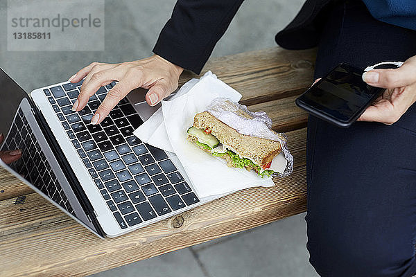 Ein Teil der Geschäftsfrau benutzt einen Laptop  während sie mit einem Smartphone und einem Sandwich an einer Bushaltestelle in der Stadt sitzt