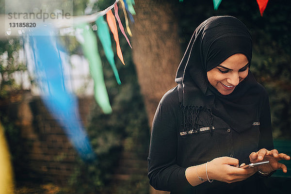 Lächelnde junge Frau im Hidschab mit Mobiltelefon im Hinterhof