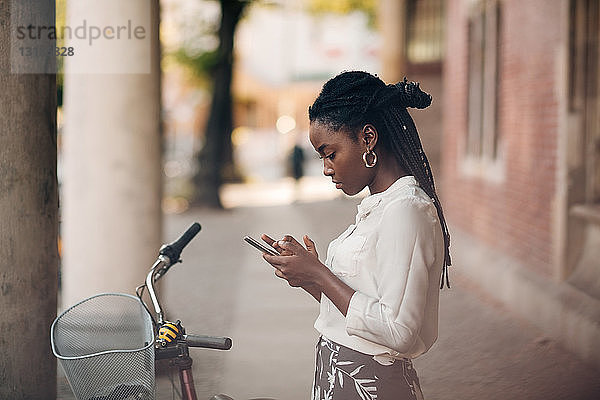 Seitenansicht einer jungen Frau  die ein Mobiltelefon benutzt  während sie mit dem Fahrrad auf einem Fußweg in der Stadt steht