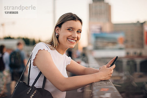 Porträt einer lächelnden jungen Frau  die ein Mobiltelefon in der Hand hält  während sie sich an ein Geländer an einer Brücke in der Stadt lehnt