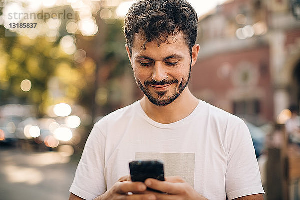 Lächelnder junger Mann benutzt Smartphone  während er in der Stadt auf der Straße steht