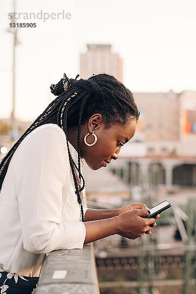 Seitenansicht einer jungen Frau  die über ein Mobiltelefon SMS-Nachrichten verschickt  während sie sich auf ein Geländer an der Brücke lehnt