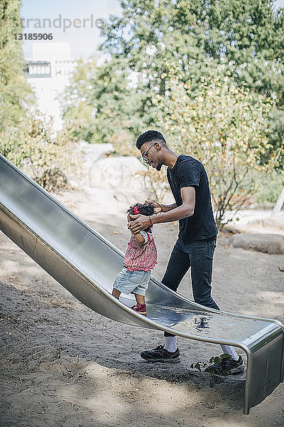 Junger Mann hält Hände einer Tochter  die auf einer Rutsche auf einem Spielplatz läuft