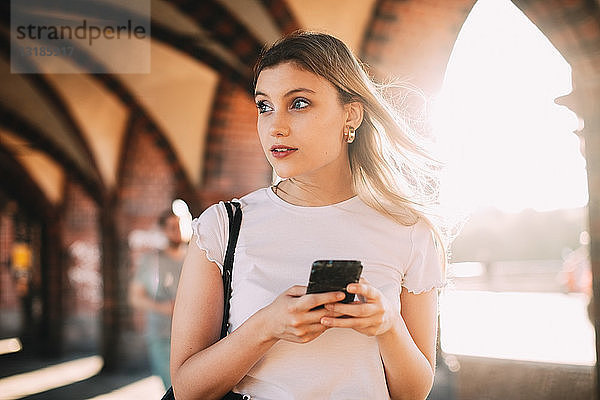 Junge Frau schaut weg  während sie auf einem Fußweg in der Stadt ein Smartphone in der Hand hält