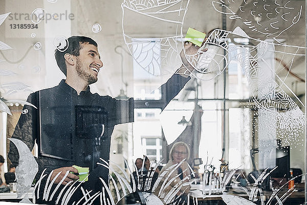 Lächelnder kreativer Geschäftsmann klebt Klebezettel auf gemustertes Glas im Büro