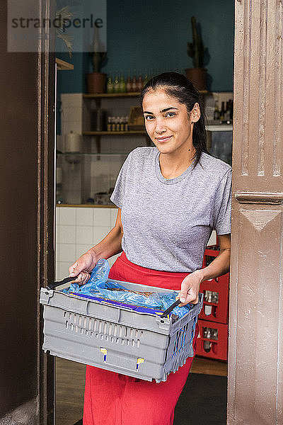 Porträt einer selbstbewussten jungen Besitzerin  die mit einer Kiste vor der Restauranttür steht