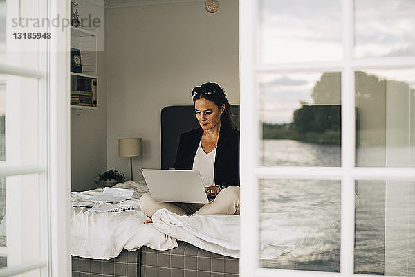 Frau mit Laptop auf dem Bett sitzend durch Türöffnung in Ferienvilla gesehen