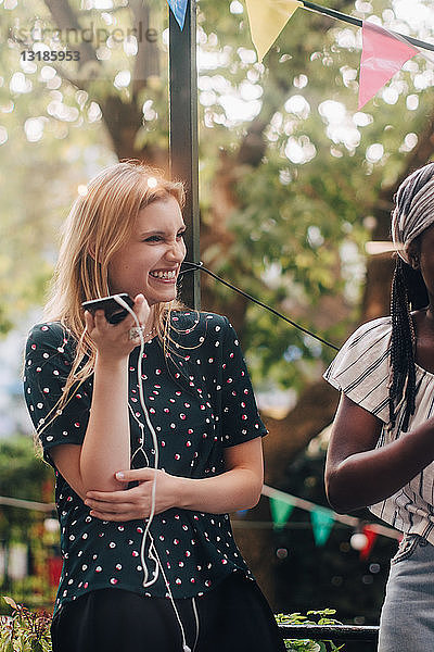 Lächelnde junge Frau hält Handy in der Hand  während sie während der Party bei einer Freundin auf dem Balkon steht