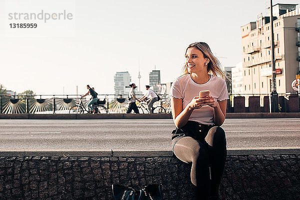 Lächelnde junge Frau hält Handy in der Hand  während sie auf einer Stützmauer an der Straße sitzt